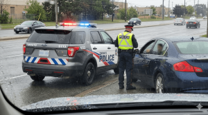 A realistic view taken through a wet windshield on a rainy day, showing a Toronto police officer conducting a traffic stop. The officer, wearing a dark uniform, a peaked cap, and a bright yellow high-visibility vest, stands at the driver's side window of a dark blue sedan. Directly behind the sedan is a dark grey Toronto Police SUV with its red and blue lights flashing. Several other cars are driving in the background on the wet, multi-lane road. This image was generated for Zamani Law.